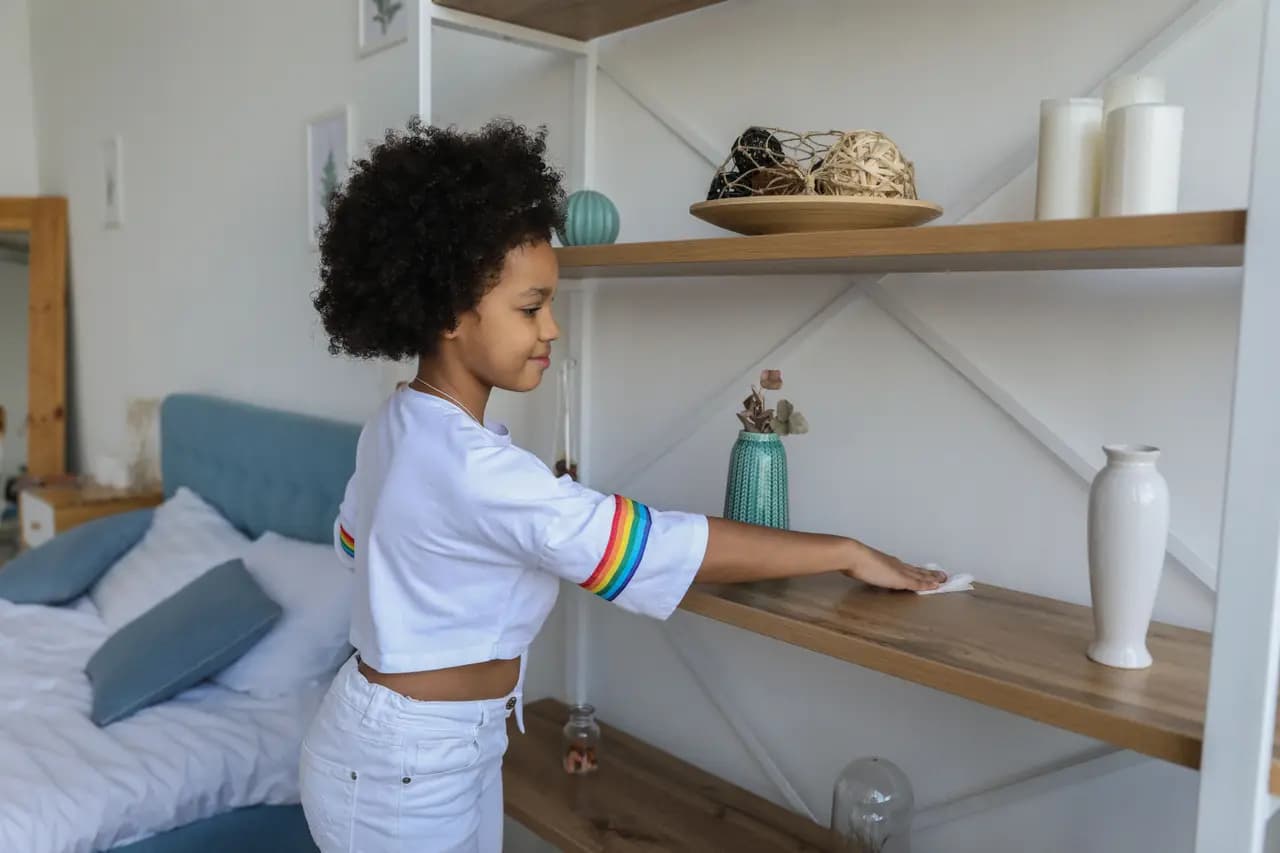 A girl dusting a shelf in a bedroom